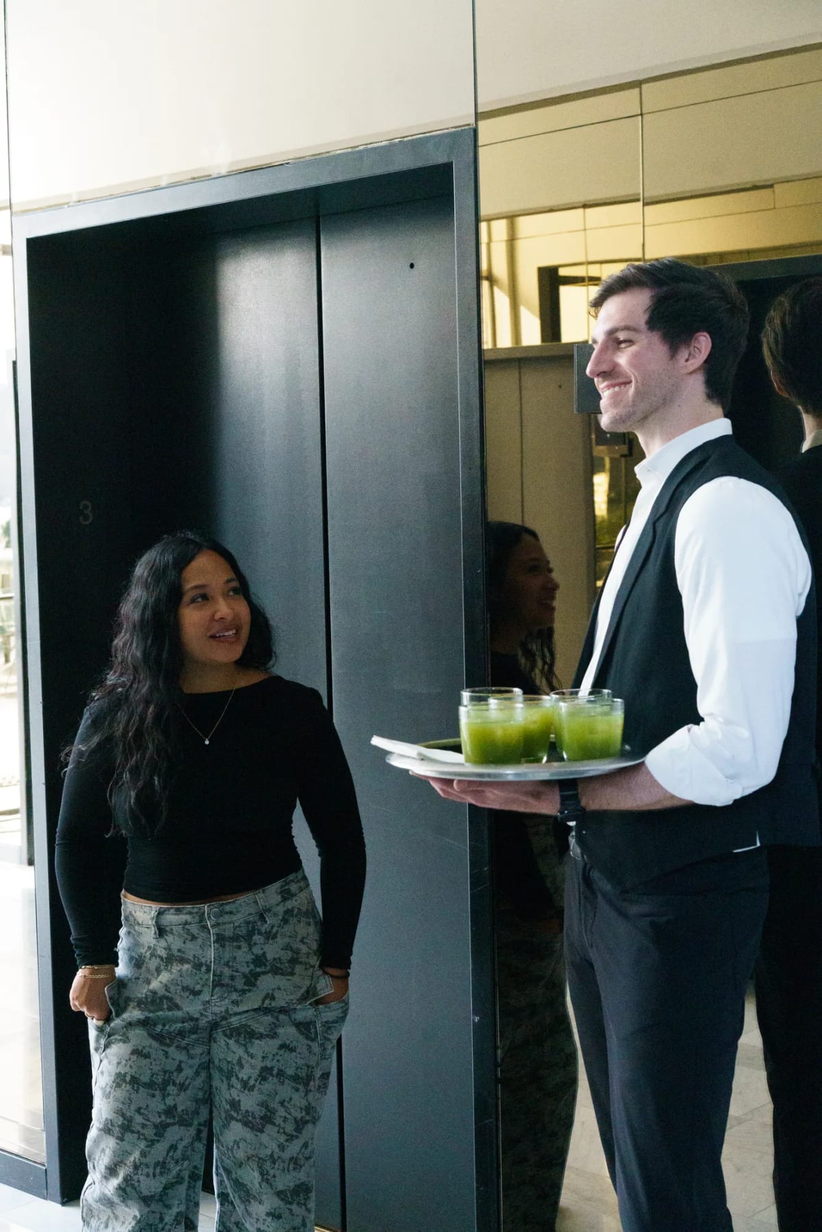A waiter offers a tray of green drinks to a guest by the elevators