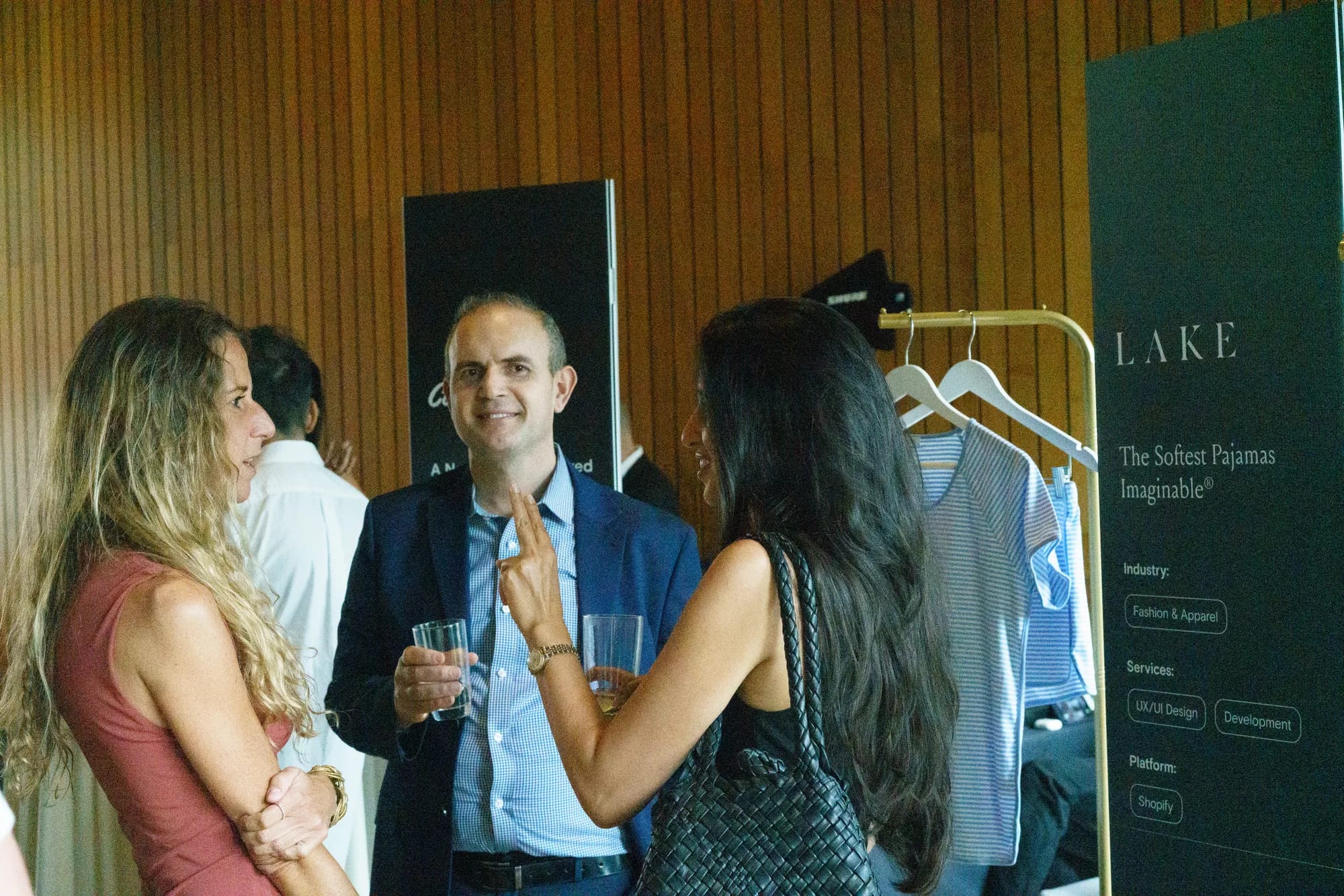 Guests chat with drinks beside a LAKE pajamas display rack