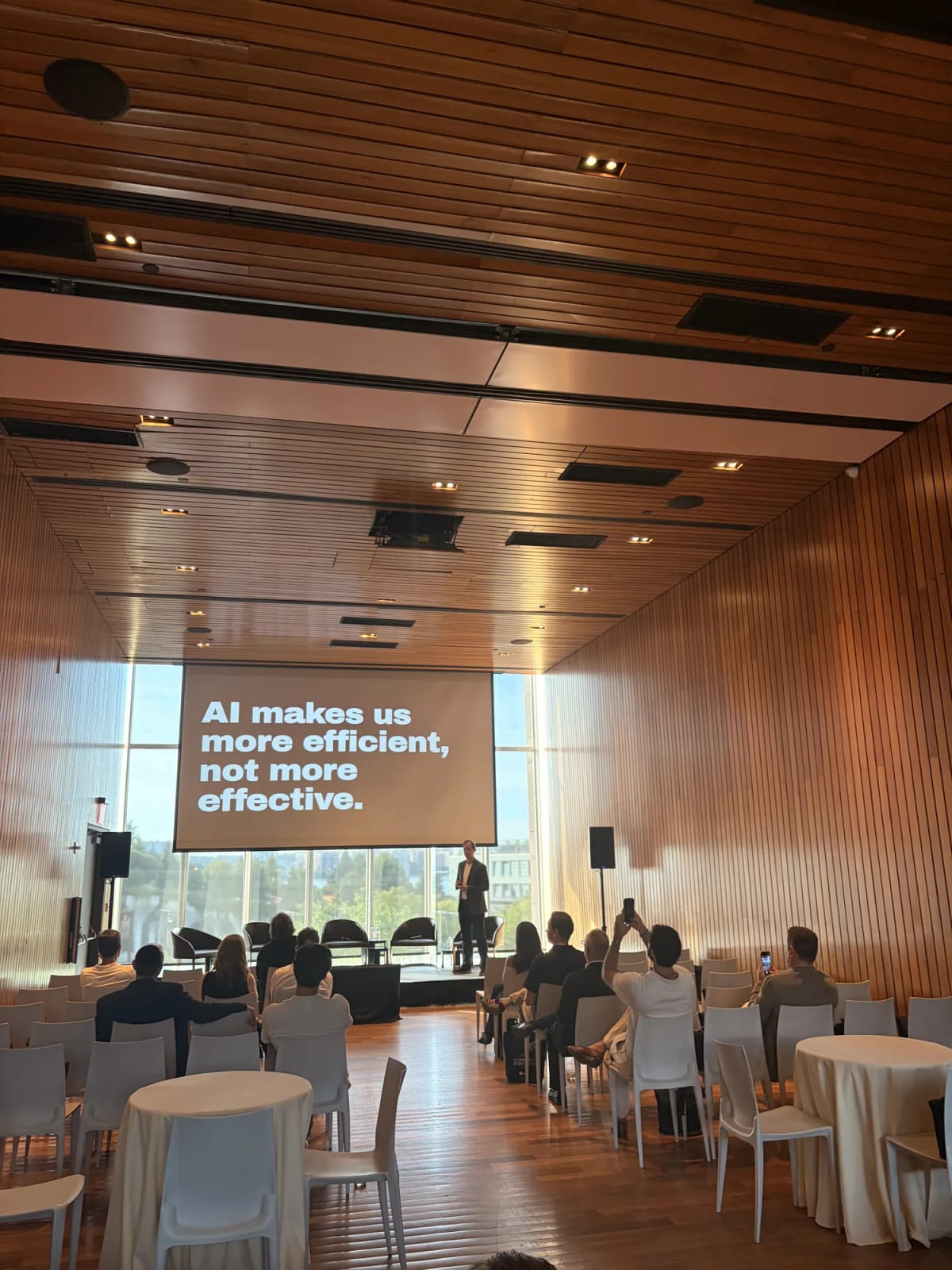 A speaker presents a slide reading "AI makes us more efficient, not more effective"