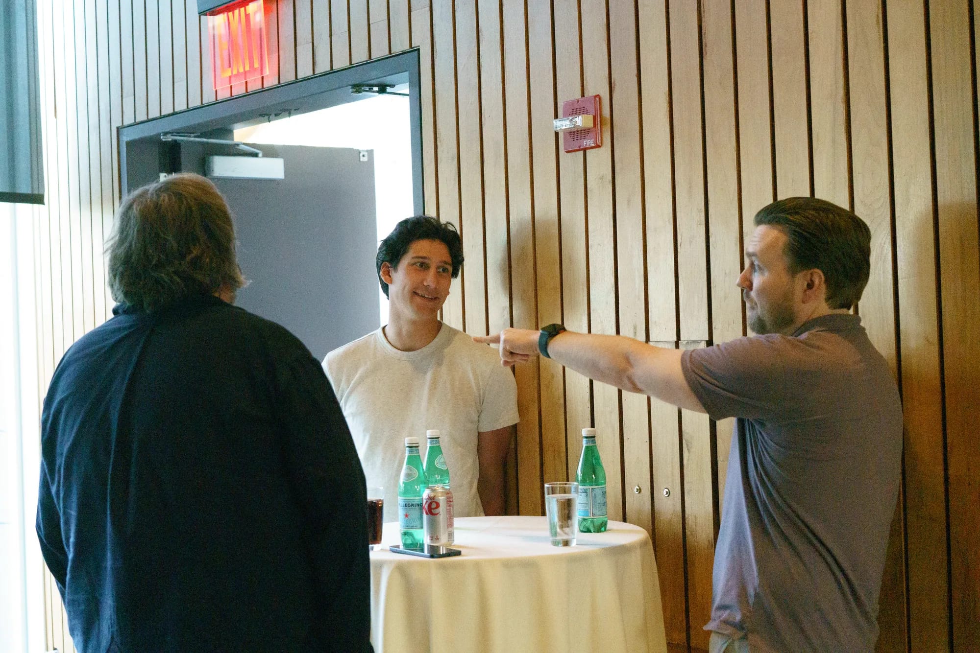 Three attendees talk around a cocktail table against a wood-paneled wall