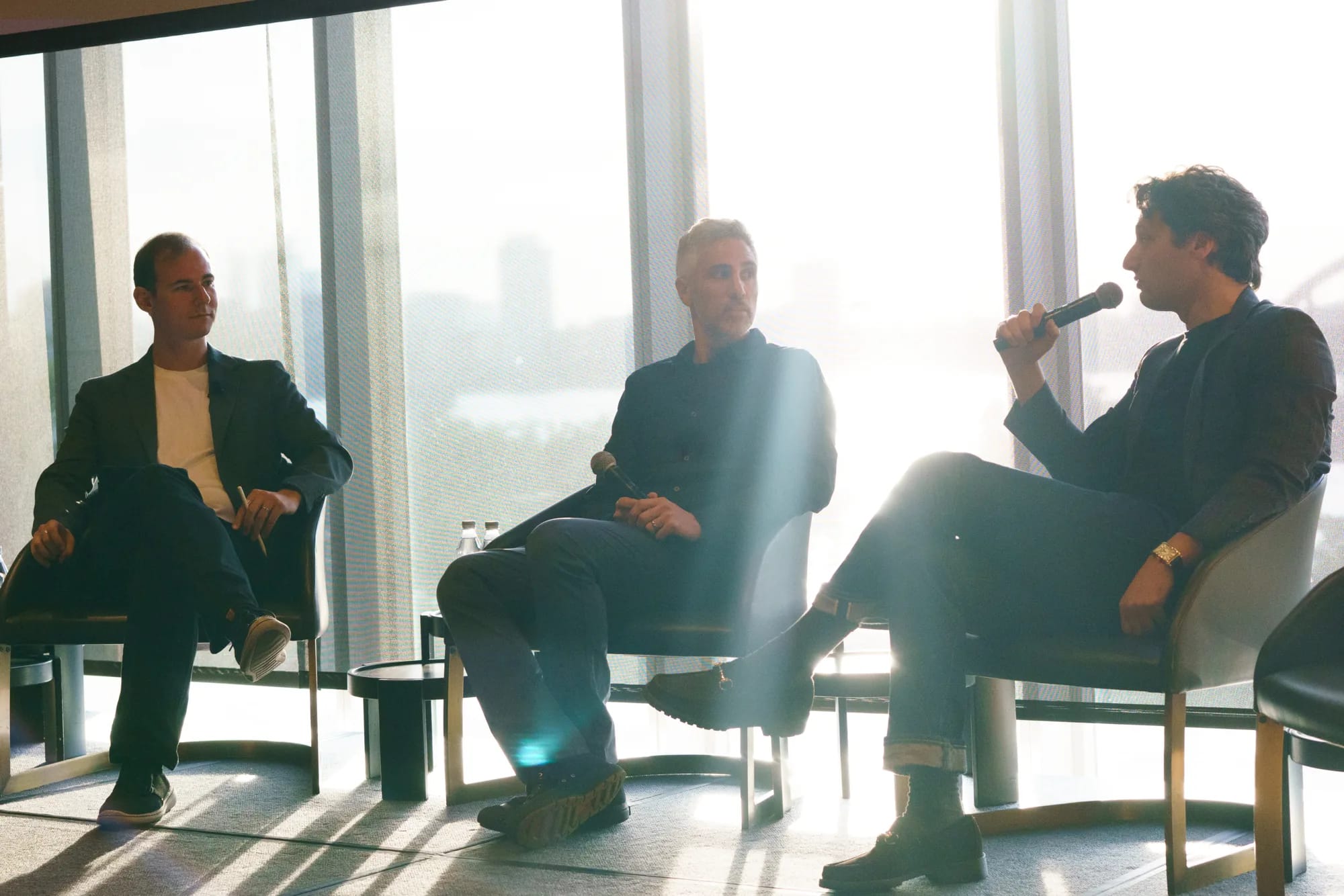 Three panelists in lounge chairs on stage, backlit by sunlit windows