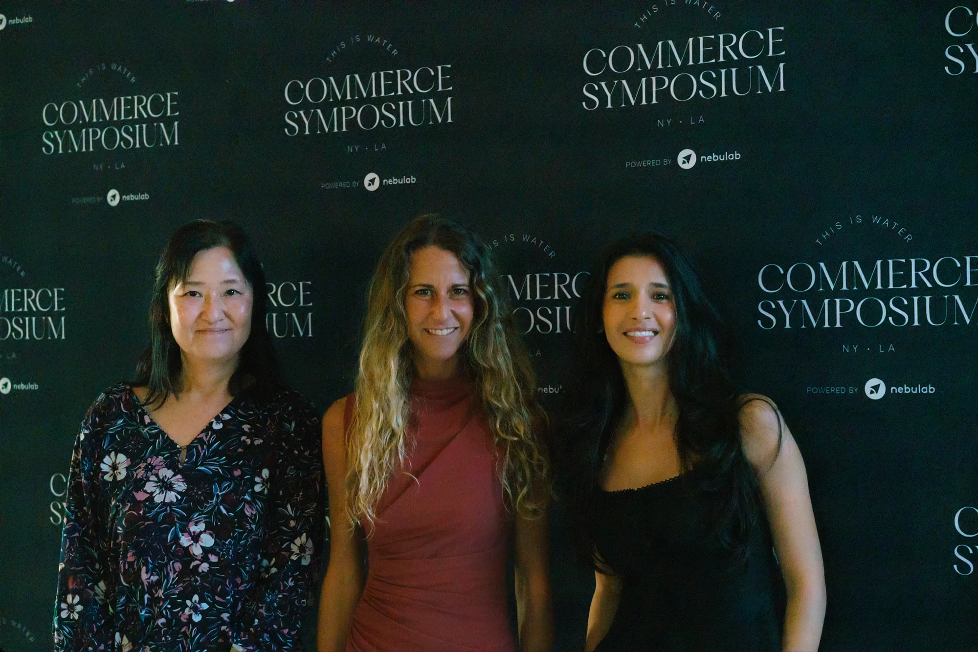 Three attendees pose in front of the Commerce Symposium step-and-repeat backdrop