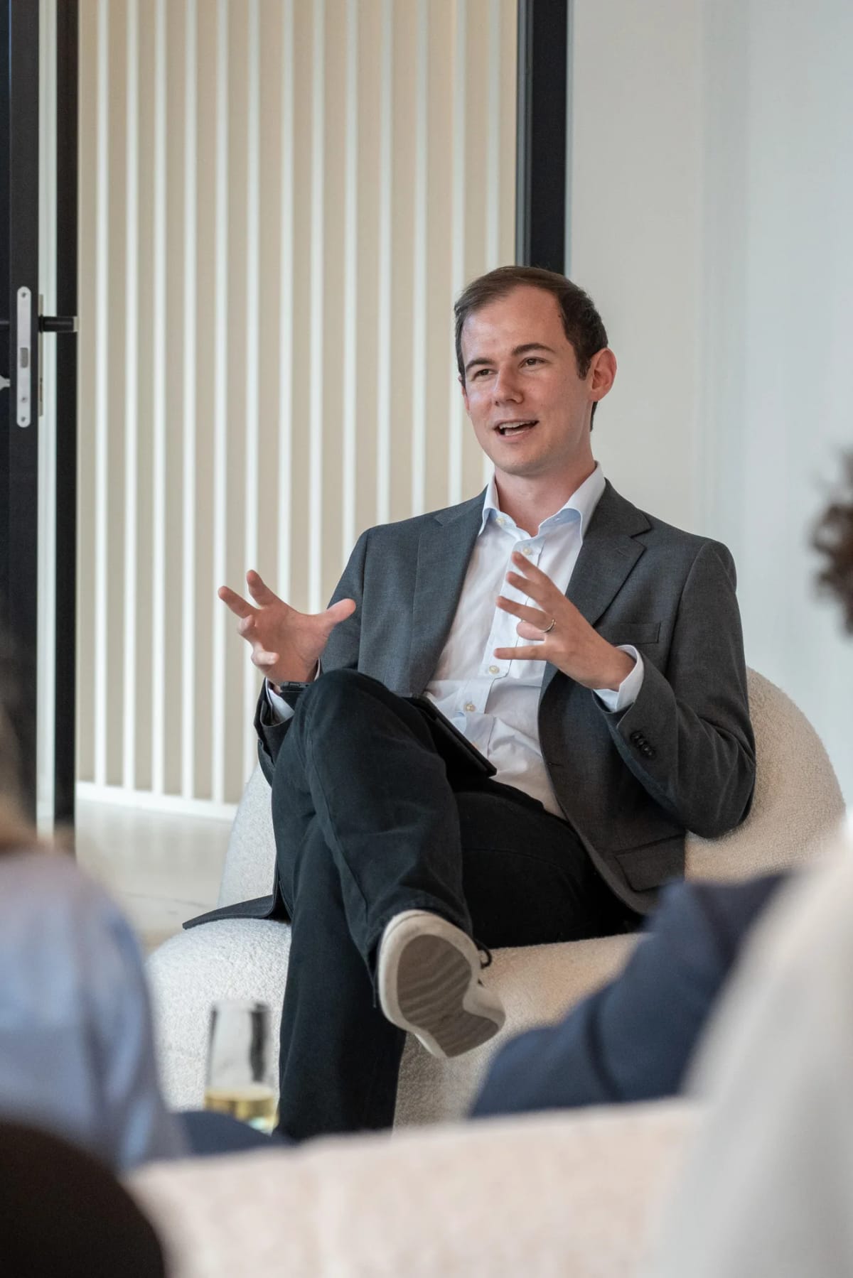 A participant in a grey blazer speaks from a white armchair