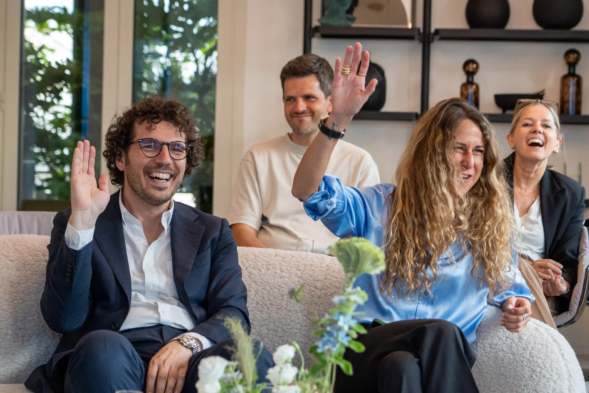 Laughing guests on a sofa raise their hands in response to a question