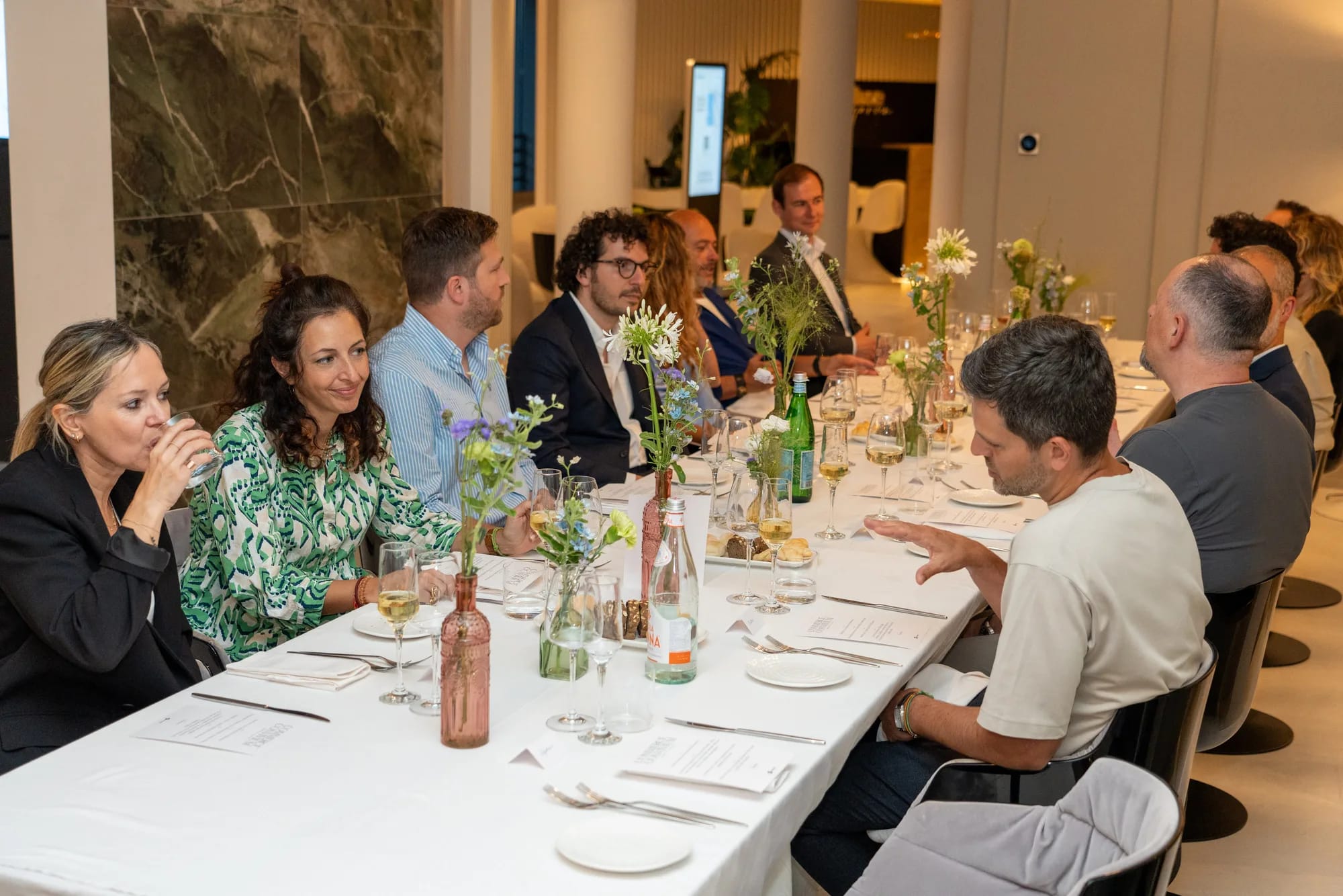 Guests talking along a long dinner table set with wine glasses and flowers