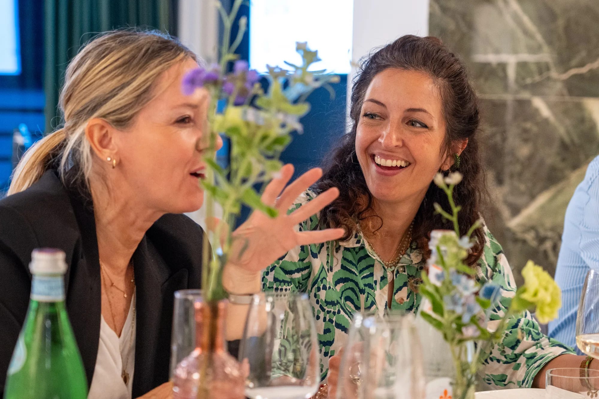 Two guests laugh together across the dinner table behind small flower vases