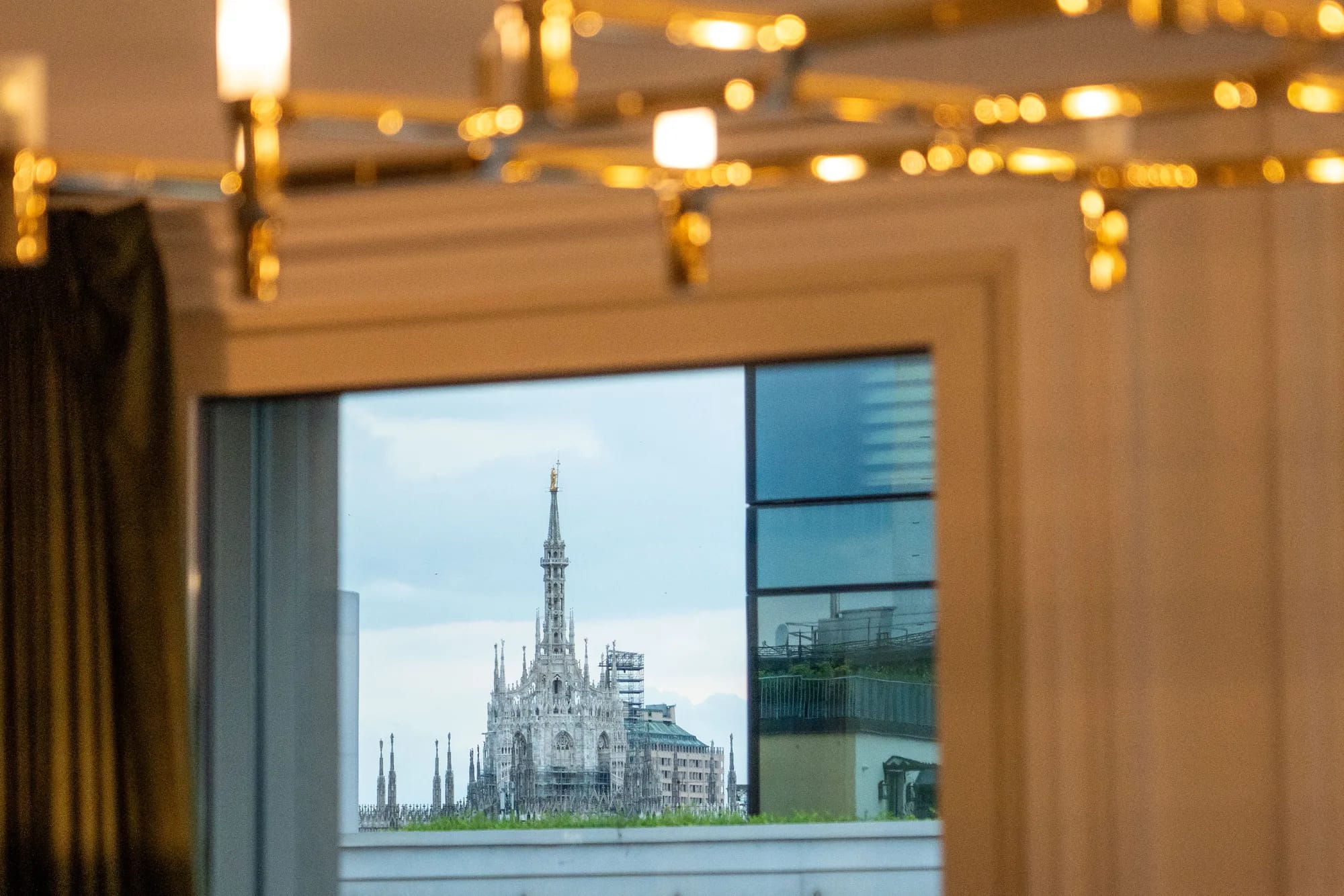 The spire of Milan Cathedral seen through a window beneath a gold chandelier