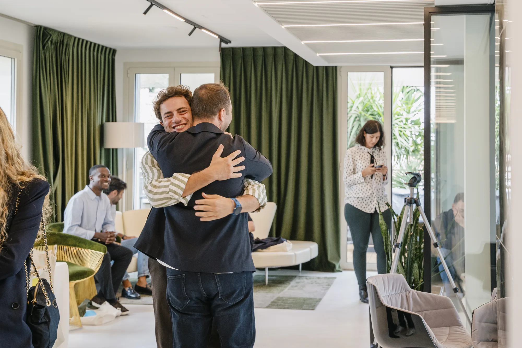 Two guests hug in a lounge with green velvet curtains as others arrive