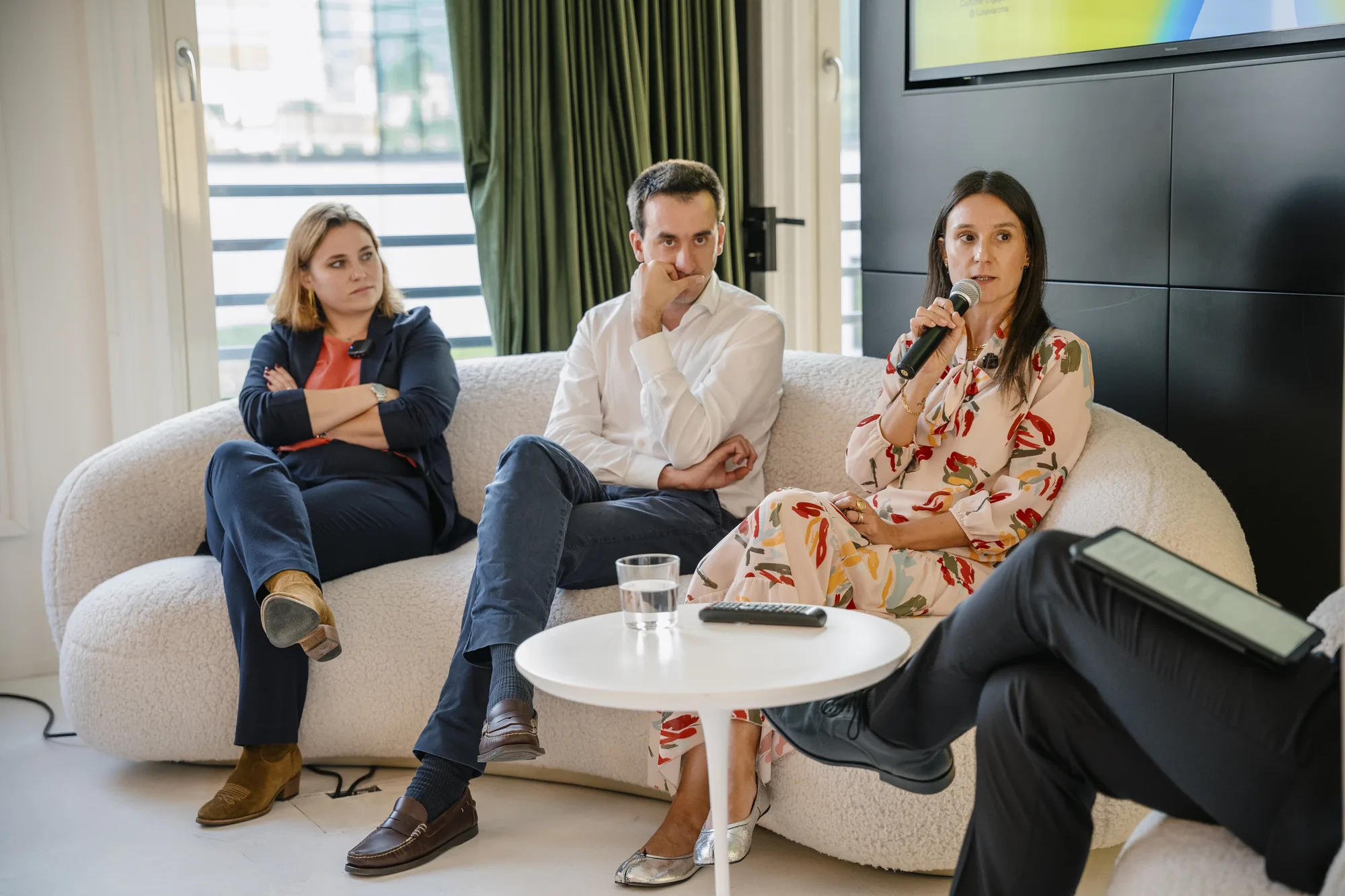 Three panelists on a sofa listen as one speaks into a microphone