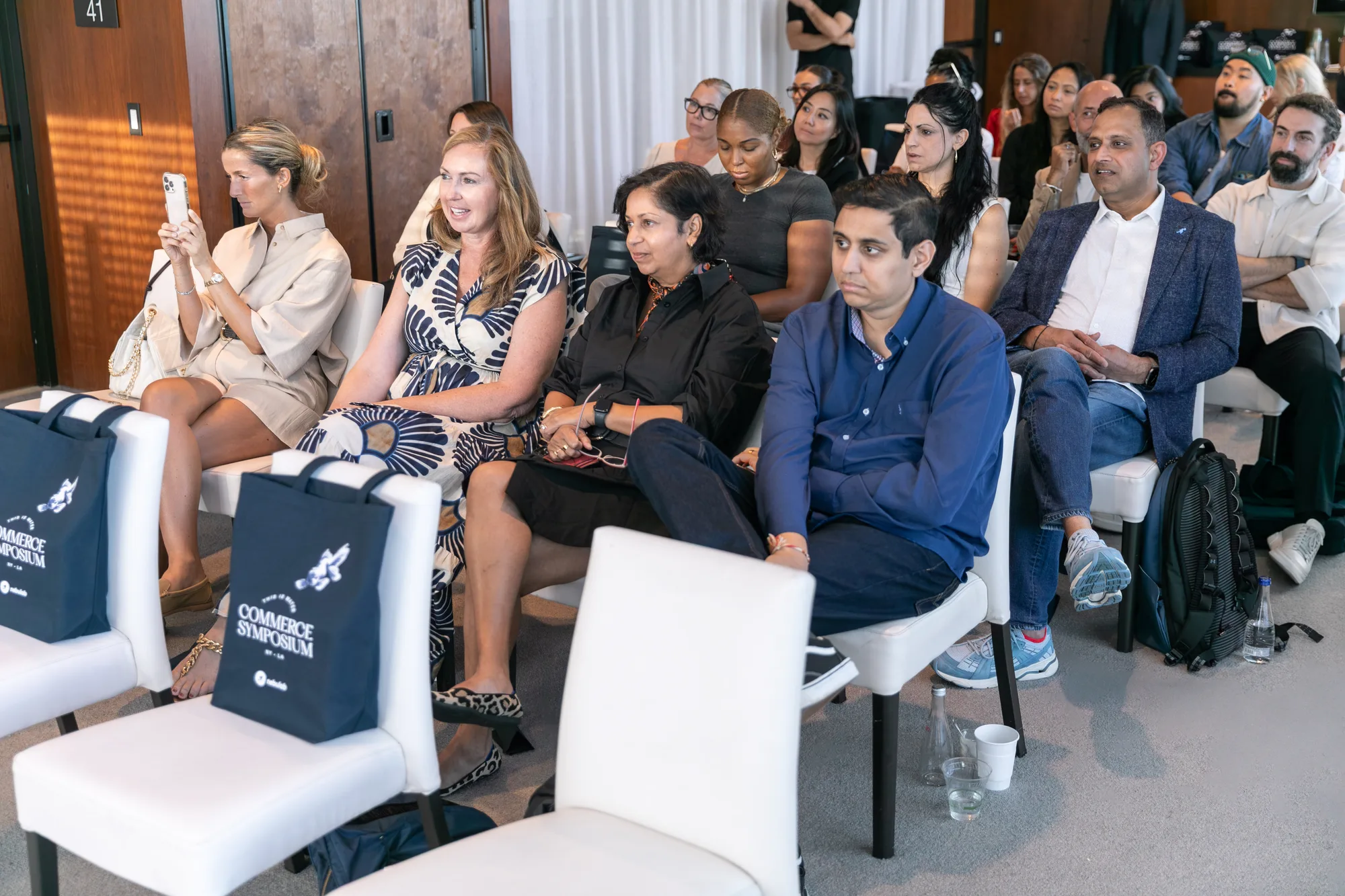 Attendees seated in rows listening, with Commerce Symposium tote bags on the chairs